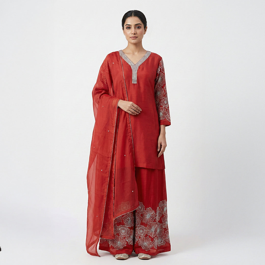 This elegant outfit is in a beautiful deep rust-red color. It features a long, simple Kurta with a sparkly silver border around the V-neckline and sleeves. It comes with matching wide-legged Palazzo pants that have large, stunning silver flower designs embroidered at the bottom. The look is completed with a matching plain Dupatta that has a thin delicate silver border, making it a classy choice for parties and family gatherings.