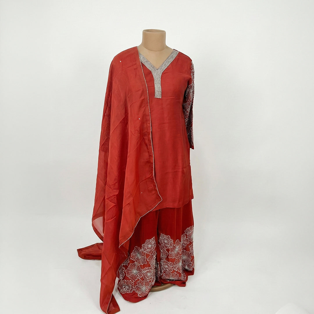This elegant outfit is in a beautiful deep rust-red color. It features a long, simple Kurta with a sparkly silver border around the V-neckline and sleeves. It comes with matching wide-legged Palazzo pants that have large, stunning silver flower designs embroidered at the bottom. The look is completed with a matching plain Dupatta that has a thin delicate silver border, making it a classy choice for parties and family gatherings.
