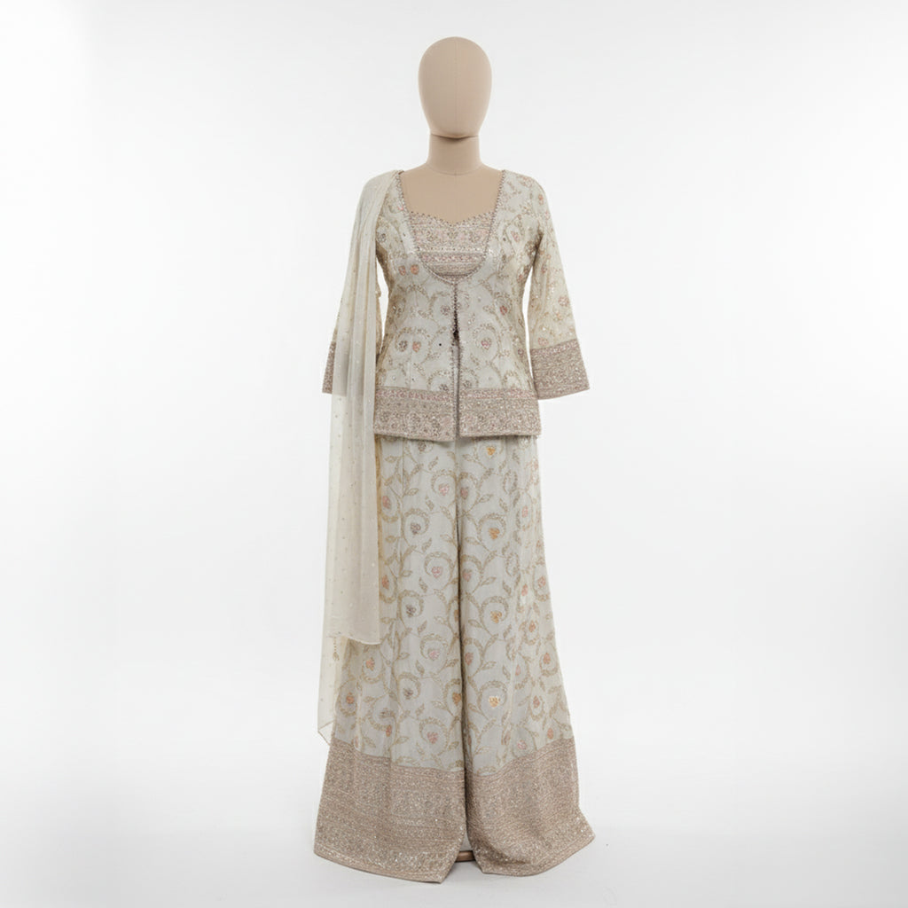 This graceful outfit is designed in a soft ivory shade with delicate golden embroidery that gives it a refined, elegant look. The fitted jacket style top has a beautiful neckline and fine handwork along the edges adding a hint of sparkle.

The wide-leg pants feature matching floral embroidery with a rich border creating a balanced and stylish appearance.


A light, sheer dupatta with tiny embroidered dots completes the outfit making it perfect for weddings, festive occasions or any event where you want to l