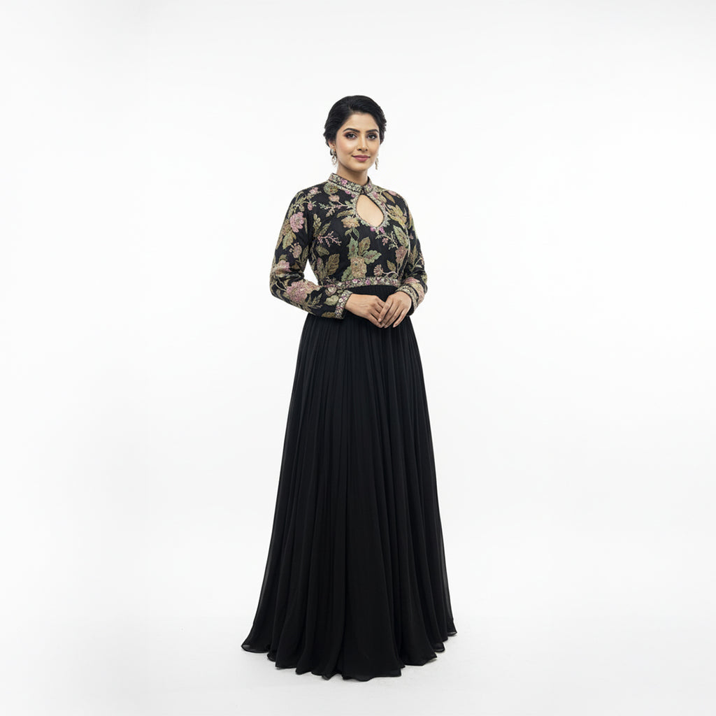 This elegant, full-length black gown features a high-neck bodice adorned with intricate, multi-colored floral embroidery.

The dress has long sleeves and a defined waistline, flowing into a voluminous, pleated skirt that reaches the floor.

The style blends traditional motifs with a modern look, making it a sophisticated choice for formal events or evening wear.
