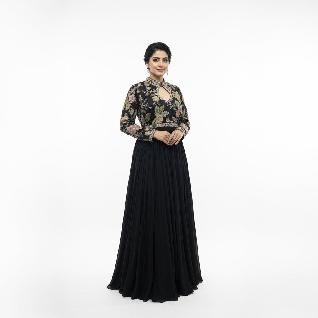 This elegant, full-length black gown features a high-neck bodice adorned with intricate, multi-colored floral embroidery.

The dress has long sleeves and a defined waistline, flowing into a voluminous, pleated skirt that reaches the floor.

The style blends traditional motifs with a modern look, making it a sophisticated choice for formal events or evening wear.