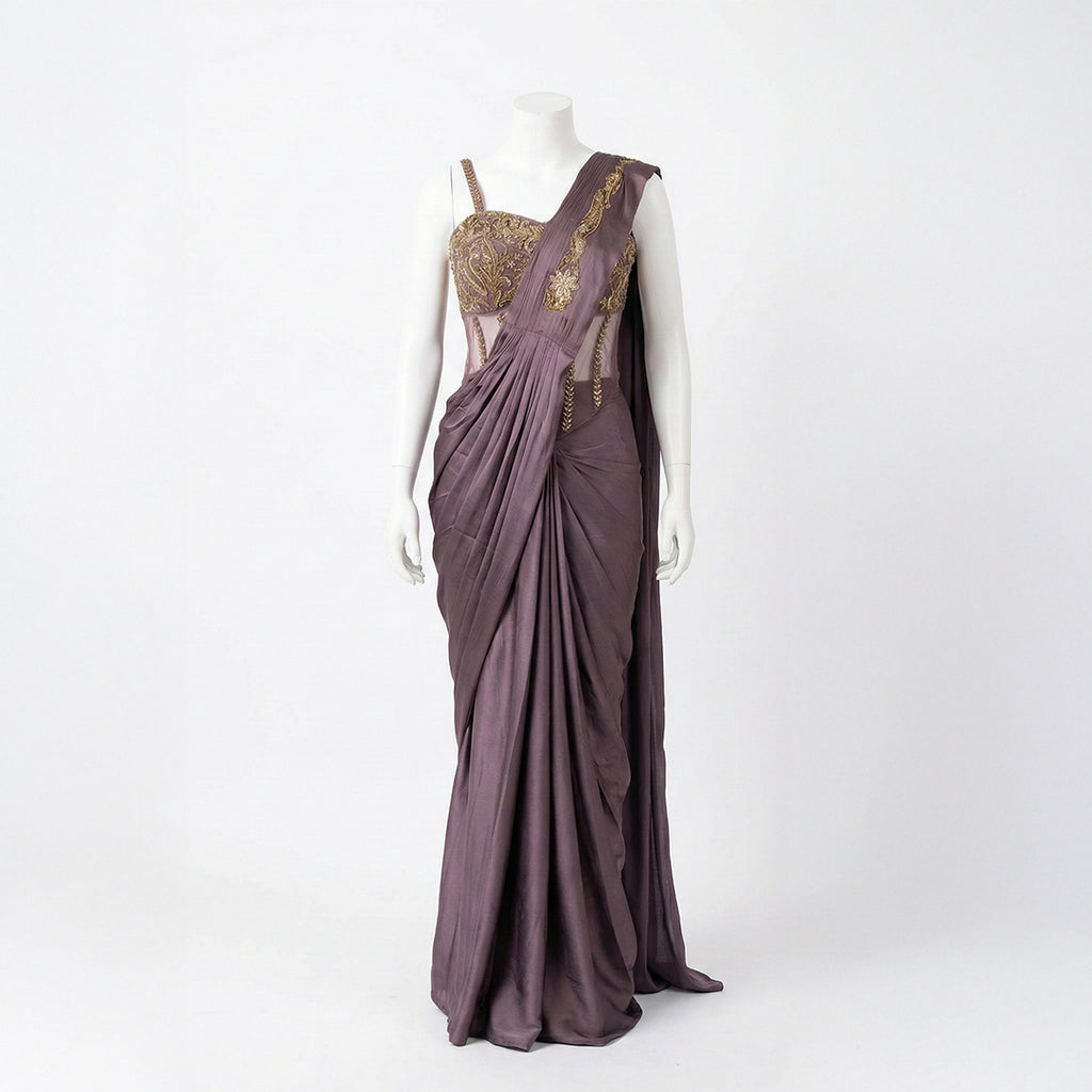 This stylish outfit is a pre-draped saree in a beautiful deep mauve color. It features a trendy corset-style blouse with thin straps and intricate gold embroidery on the upper part. The saree is draped perfectly with neat pleats in the front, and the fabric feels smooth and silky. The look is completed with an embroidered border on the shoulder drape, making it a modern and elegant choice for cocktail parties.