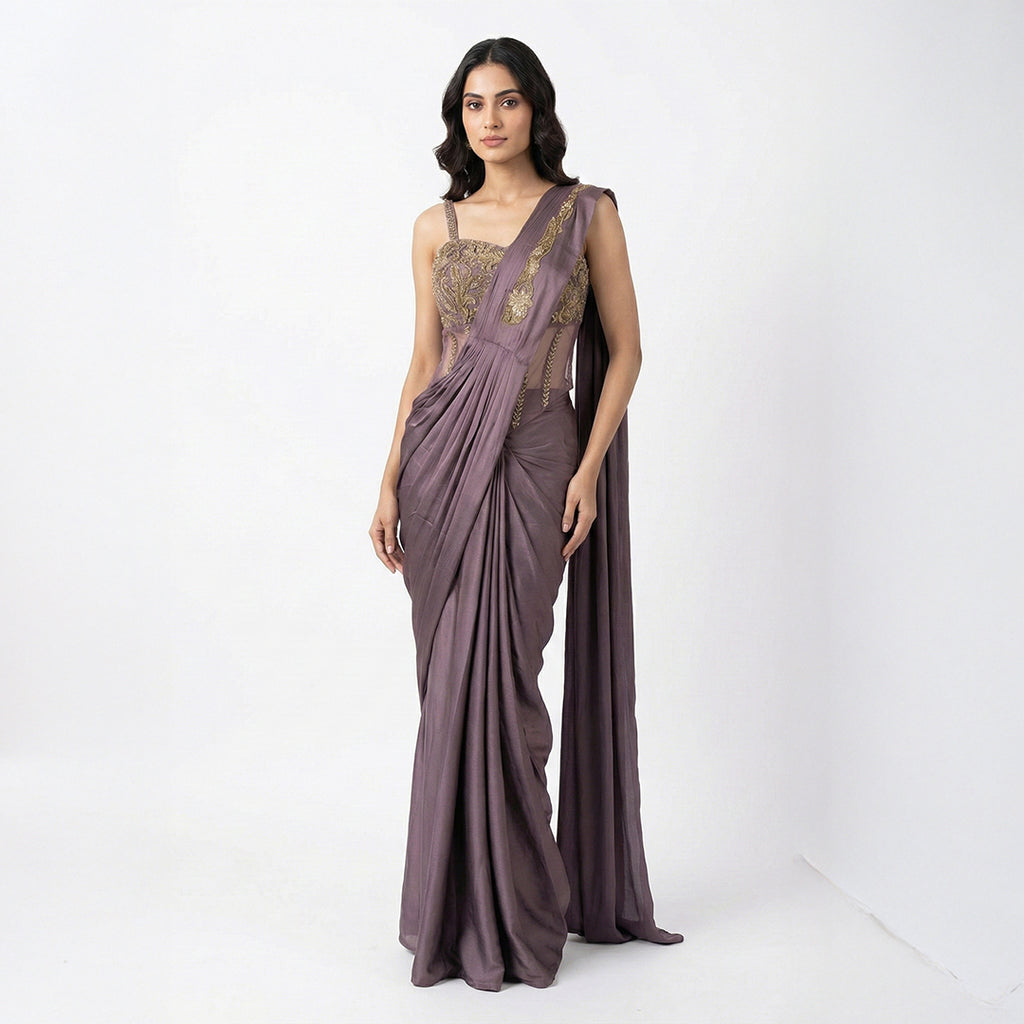 This stylish outfit is a pre-draped saree in a beautiful deep mauve color. It features a trendy corset-style blouse with thin straps and intricate gold embroidery on the upper part. The saree is draped perfectly with neat pleats in the front, and the fabric feels smooth and silky. The look is completed with an embroidered border on the shoulder drape, making it a modern and elegant choice for cocktail parties.