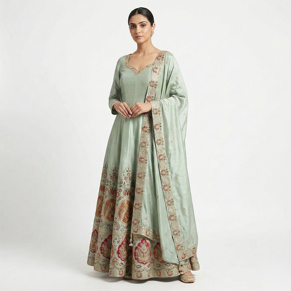 This elegant outfit is in a shiny sea-green color. It features a long, flowy Anarkali kurta with long sleeves and delicate work around the neckline. The bottom of the dress is heavily decorated with colorful red and gold floral embroidery. It comes with matching pants that fit comfortably. The look is finished with a matching Dupatta that has a beautiful embroidered border, making it perfect for weddings and festivals.